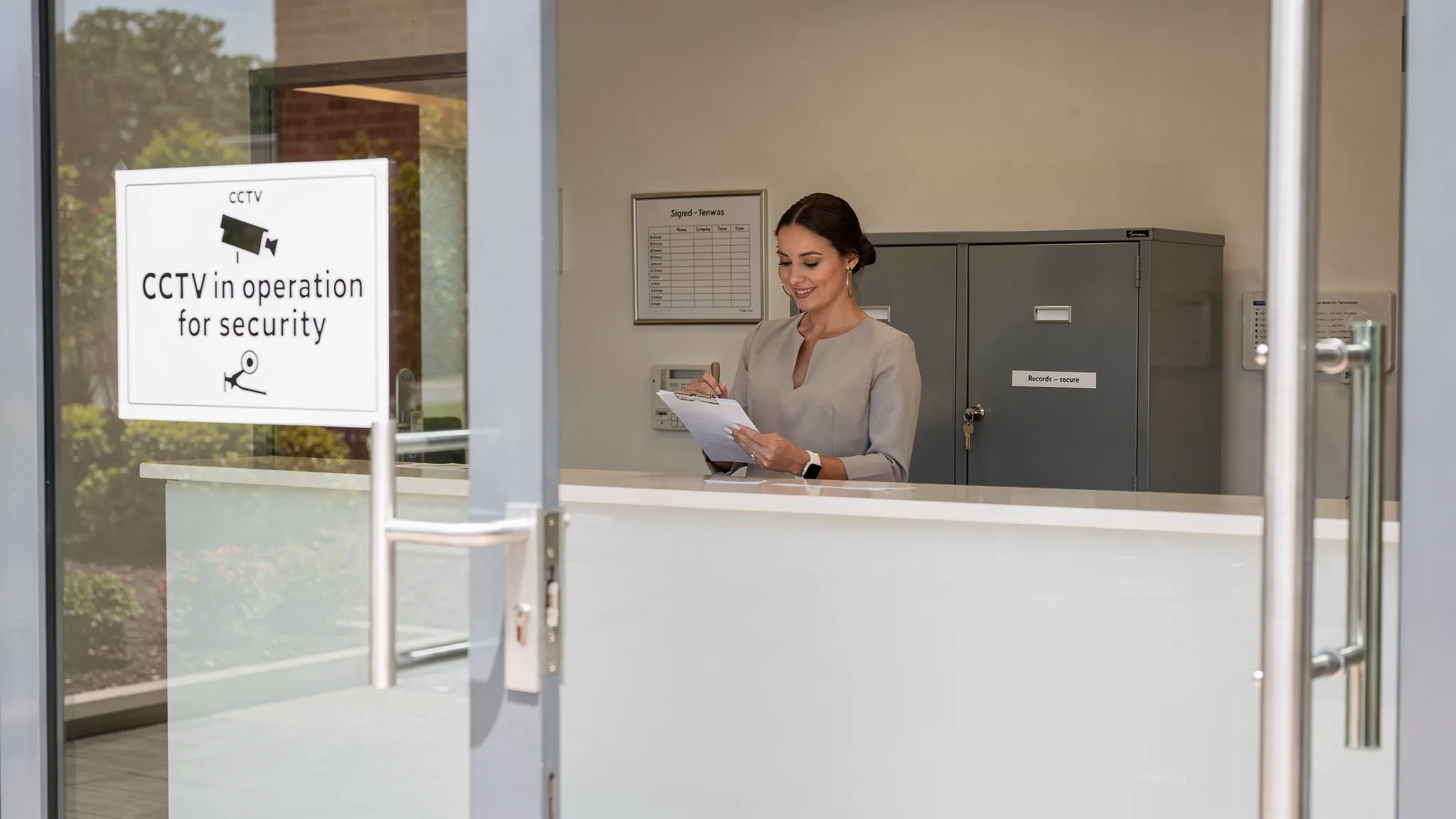 An office reception scene showing a CCTV sign at the entrance, a staff member handling a visitor sign-in sheet, and a secure locked cabinet behind the desk. The image highlights good practice: clear signage, minimal collection fields on the sign-in s...
